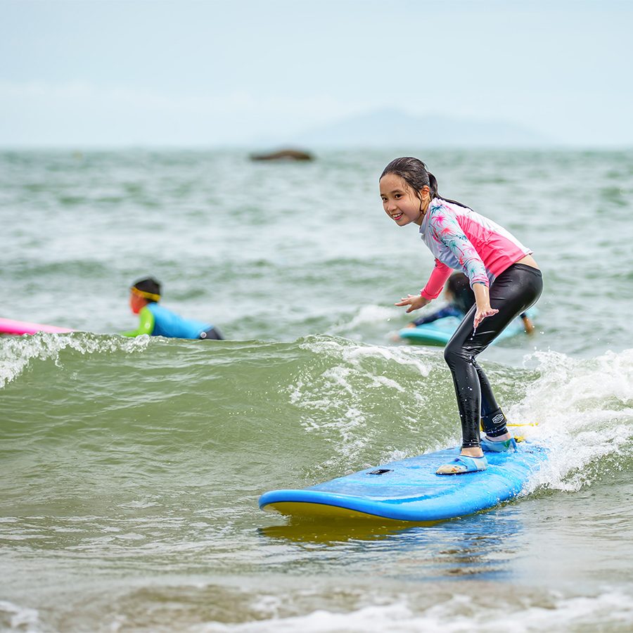 A girl surfing over a wave while onlookers watch in the background. 